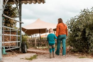 Een jongetje met een broek vol zand en een volwassen vrouw lopen hand in hand over het terrein van een kampeerboerderij