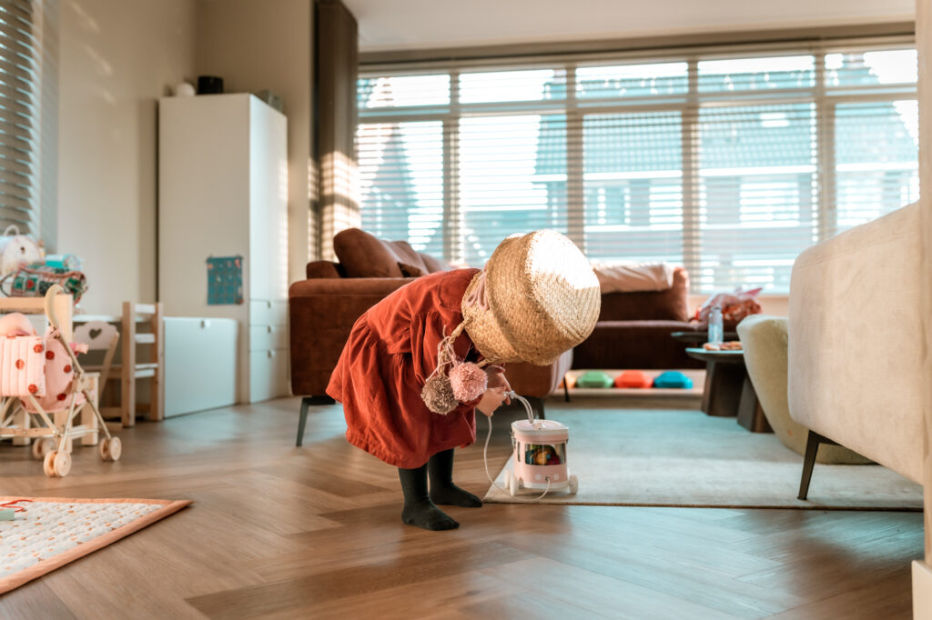 Meisje in rode jurk staat midden in de woonkamer met een rieten speelmand op haar hoofd. Ze heeft het touw van een trekfiguur vast. Award winnende foto bij This is reportage family
