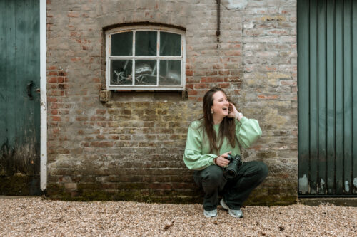 Vrouw zit op haar hurken voor een oud stenen gebouw met een systeemcamera in haar hand. Ze lacht en kijkt naar links terwijl ze haar handen door haar haar haalt. Foto genomen voor Moment Fotografie
