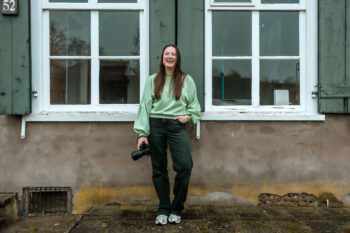 Vrouw staat voor oud gebouw met houten luiken en lacht voluit. Ze heeft een systeemcamera in haar hand
