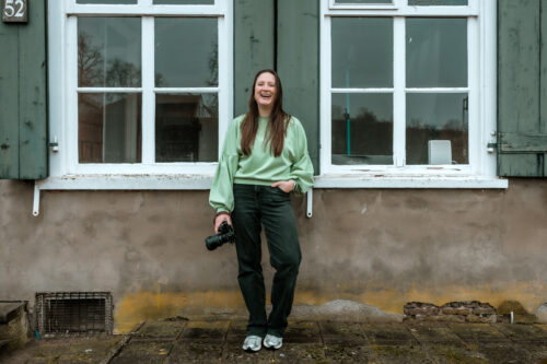 Vrouw staat voor oud gebouw met houten luiken en lacht voluit. Ze heeft een systeemcamera in haar hand