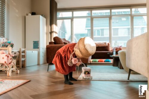 Meisje in rode jurk staat midden in de woonkamer met een rieten speelmand op haar hoofd. Ze heeft het touw van een trekfiguur vast. Award winnende foto bij This is reportage family